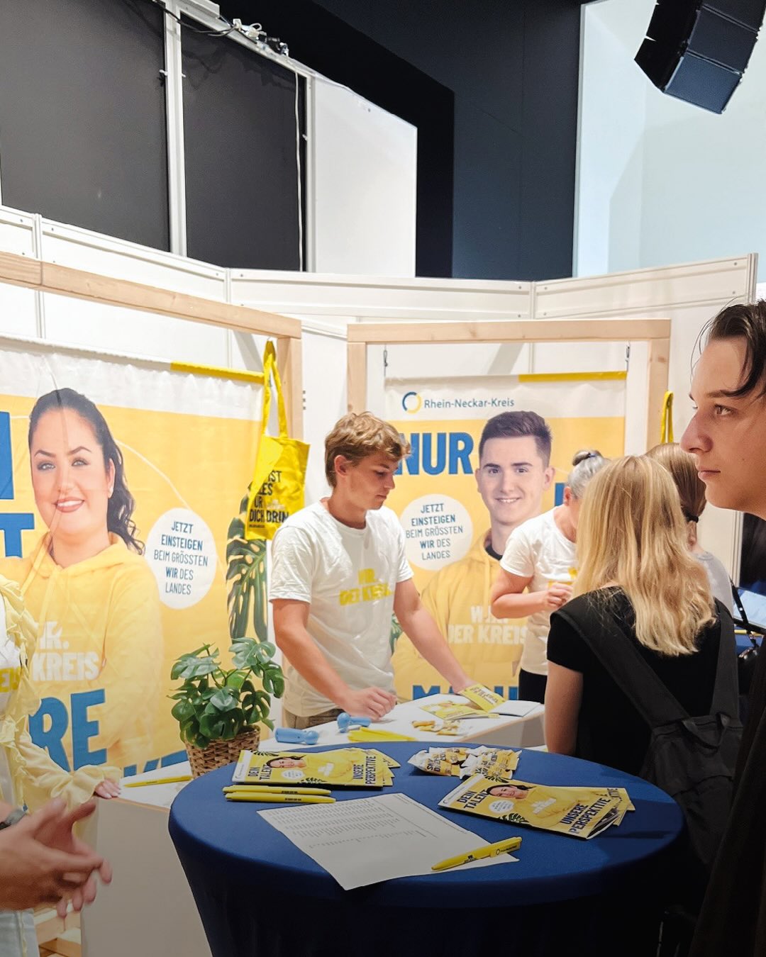 Messestand des Rhein-Neckar-Kreises mit gelben Bannern, auf denen „Rhein-Neckar-Kreis“ und „Jetzt einsteigen beim größten Wir des Landes“ steht. Mehrere Personen stehen um einen runden Tisch mit blauer Tischdecke, auf dem Broschüren, Stifte und ein Dokument liegen. Eine Person in weißem T-Shirt sortiert Materialien auf dem Tisch, während andere Personen im Vordergrund und Hintergrund stehen. (KI-Alternativtext)