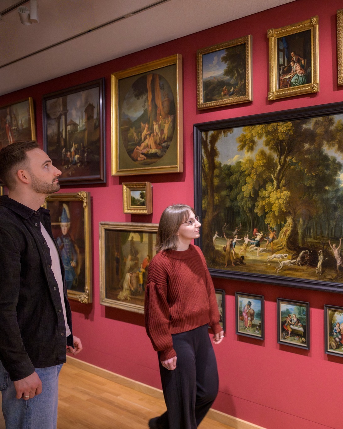 Dieser Alternativtext wurde KI-generiert: Zwei Personen betrachten Gemälde in einem Ausstellungsraum mit roten Wänden. Mehrere gerahmte Kunstwerke hängen an den Wänden.