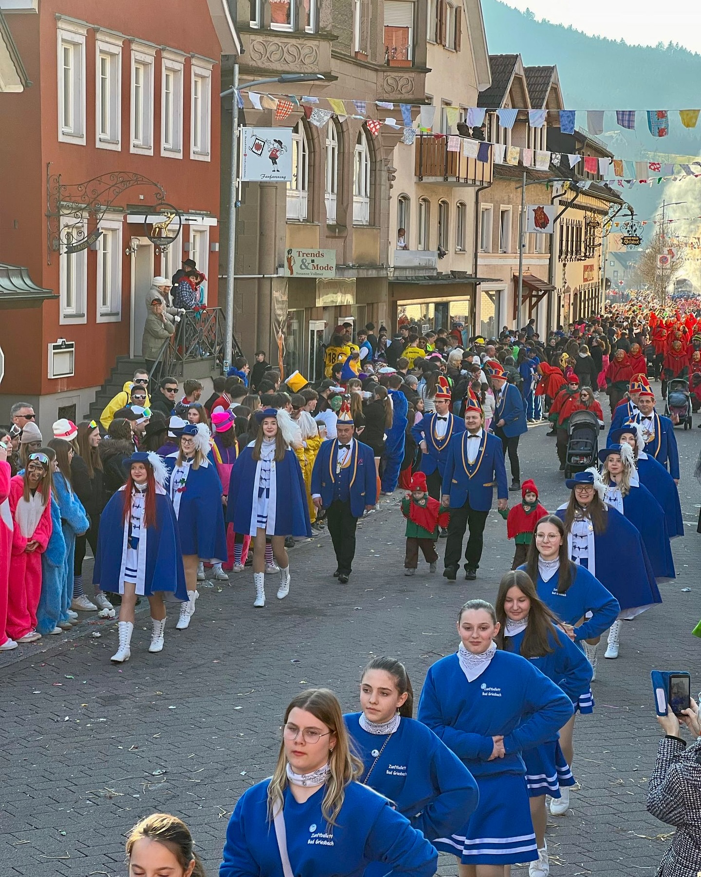 Das Foto zeigt eine Parade auf einer belebten Straße flankiert von Häusern. Die Teilnehmenden tragen Kostümierungen in Blautönen, weiße Stiefel und Mützen.
