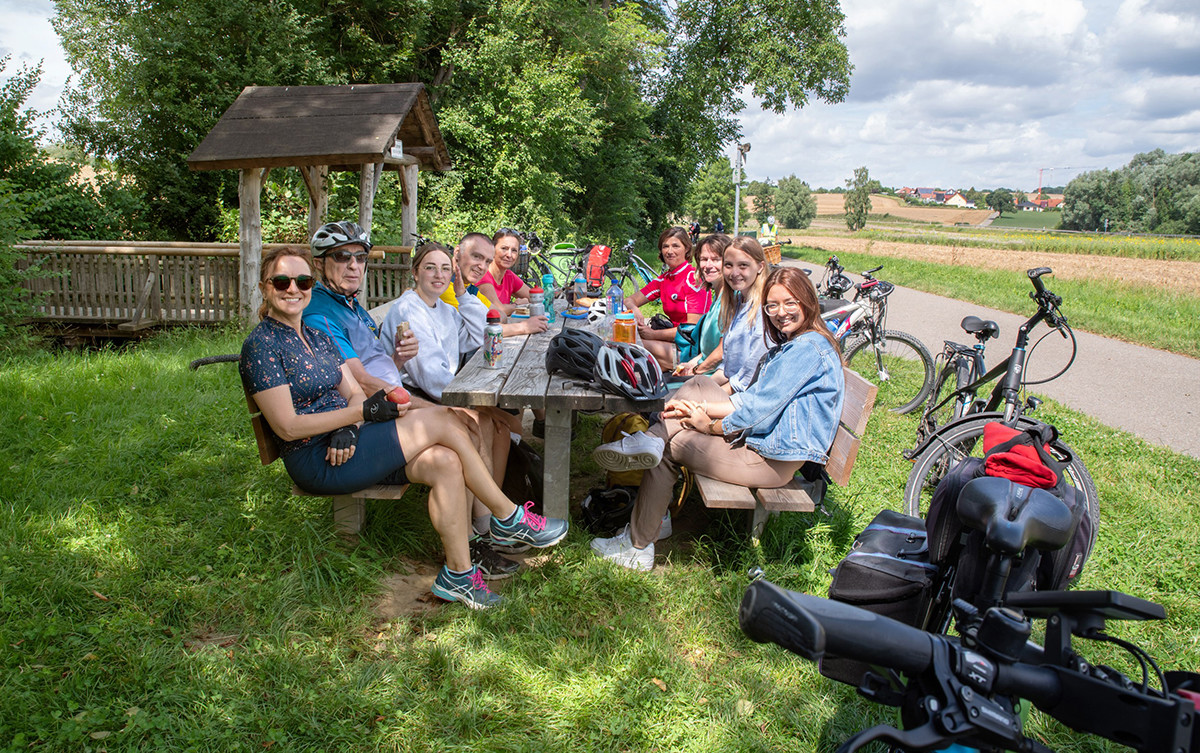 Eine Gruppe von Personen sitzt an einem Holztisch im Freien und macht eine Pause bei einer Radtour. Mehrere Fahrräder stehen im Gras, auf dem Tisch liegen Helme und Getränke. (KI-Alternativtext)