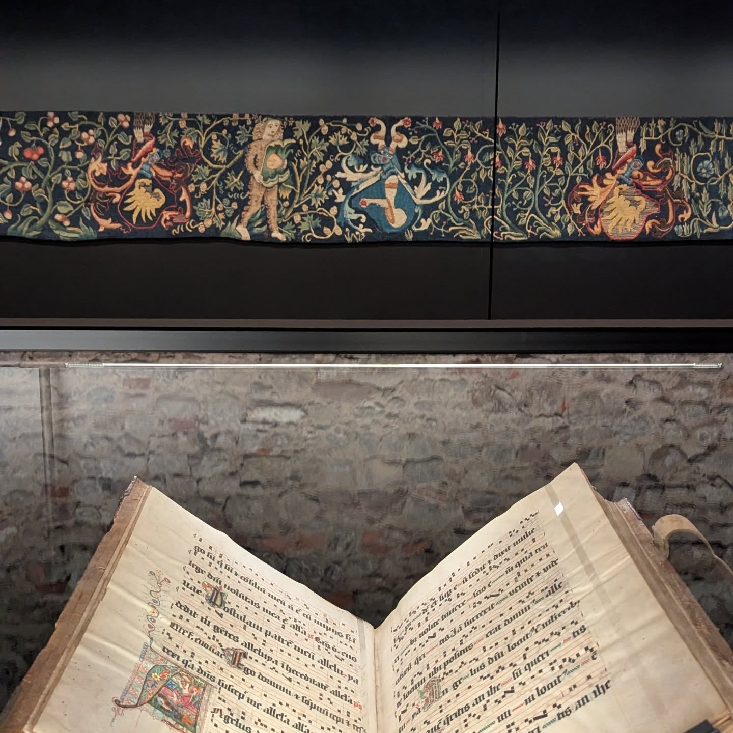 Dieser Alternativtext wurde KI-generiert: Ein Innenraum zeigt eine steinernen Wand und einen geöffneten, alten Folianten. Darüber hängt ein farbenfroher Wandteppich mit Figuren und Schriftzeichen.