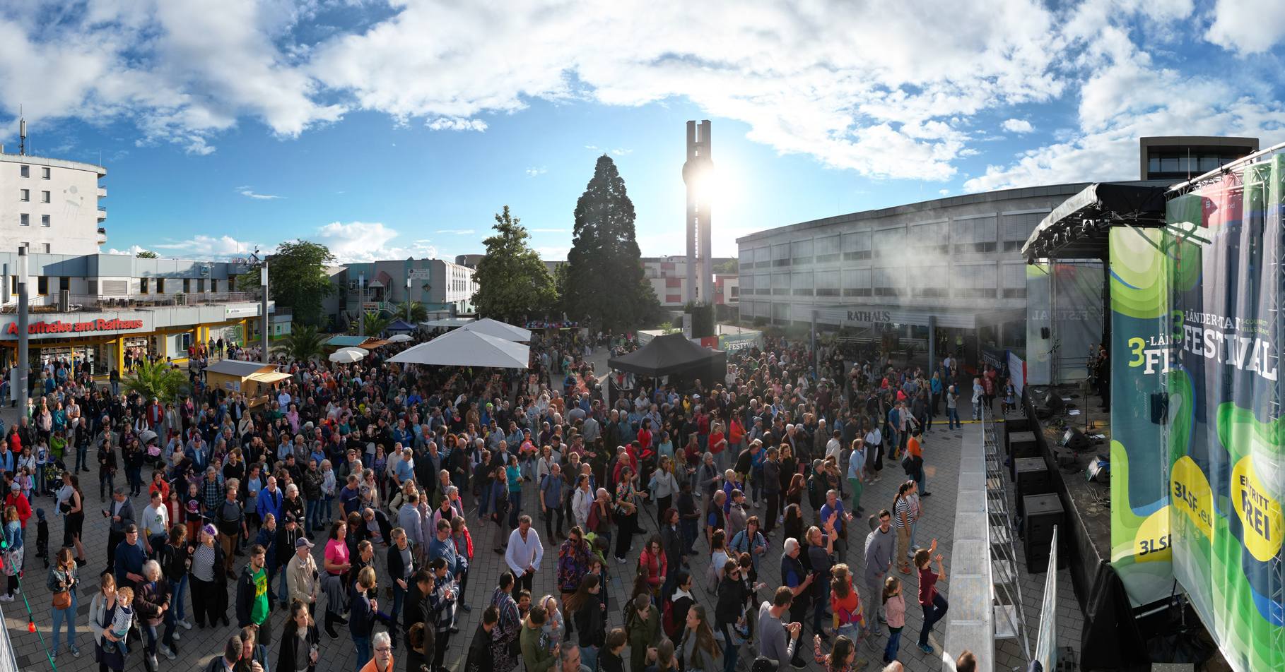 Ein großes Fest mit Musik und vielen Menschen findet im Freien statt.  Auf einem großen Veranstaltungsort stehen mehrere Bühnen.
