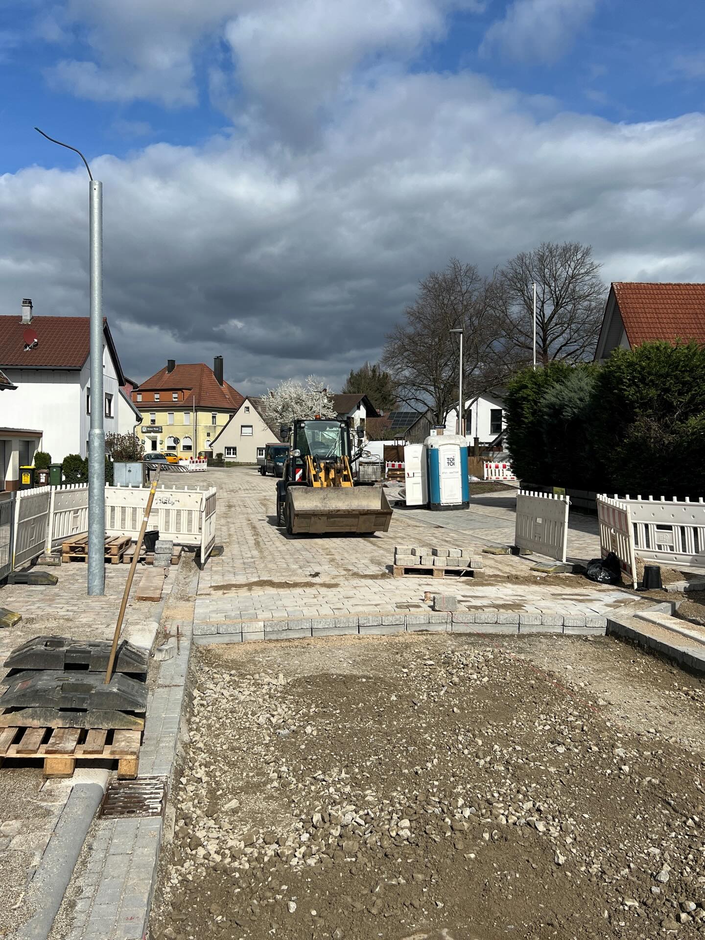 Dieser Alternativtext wurde KI-generiert: Eine Straße mit Bauarbeiten. Im Hintergrund sind Häuser und ein Baum unter einem bewölkten Himmel zu sehen.