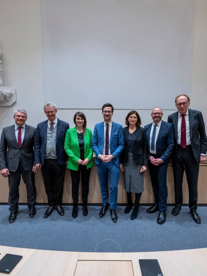 Dieser Alternativtext wurde KI-generiert: Eine Gruppe von Personen in formeller Kleidung steht vor einer hellen Wand und einer Bühne. Das Bild ist farbig und zeigt eine symmetrische Anordnung der Personen.