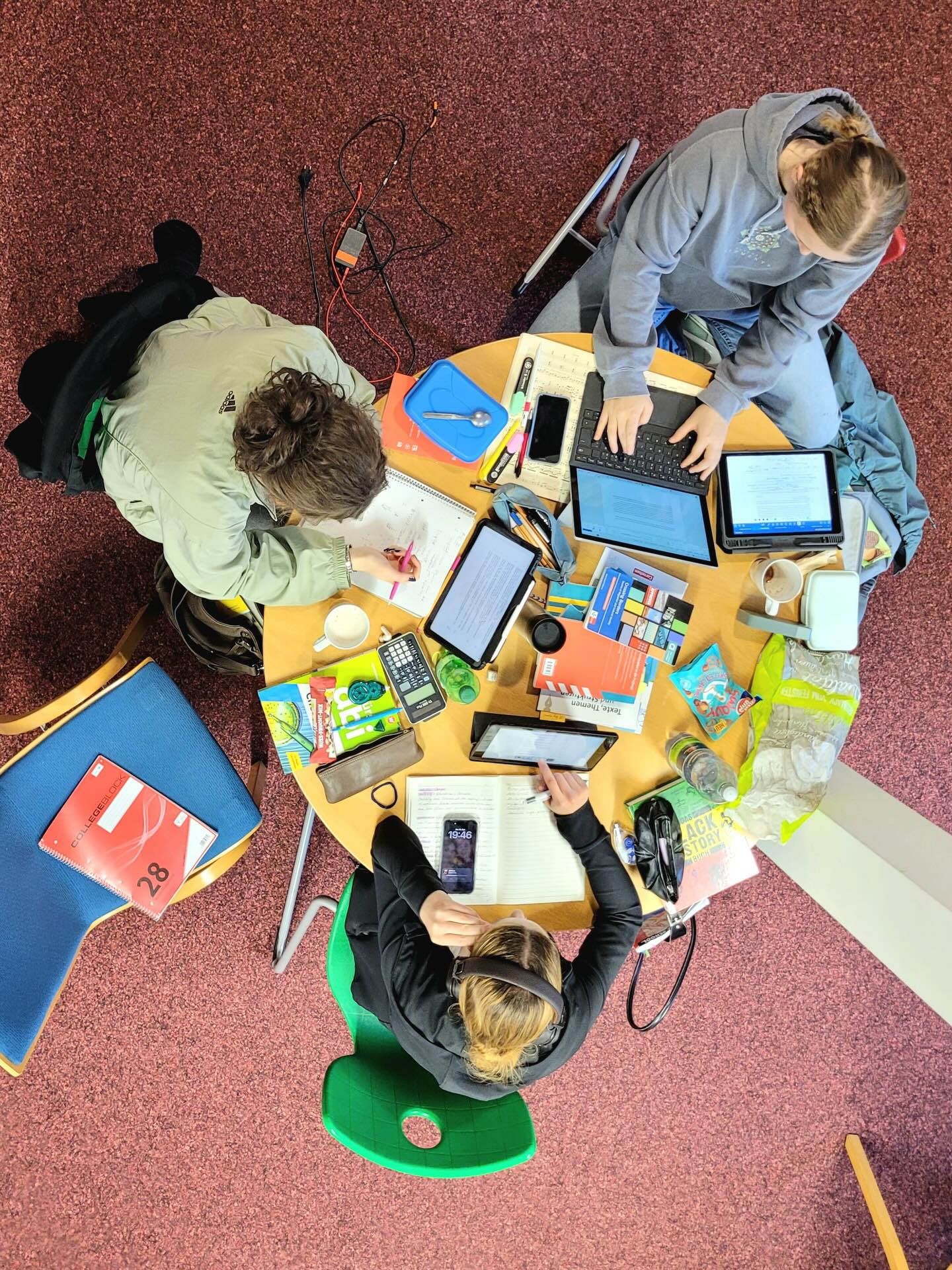 Dieser Alternativtext wurde KI-generiert: Ein farbiges Bild zeigt eine Vogelperspektive auf eine Gruppe von Personen, die um einen Tisch sitzen und verschiedene Gegenstände wie Bücher, Tablets und Schreibutensilien benutzen. Der Tisch ist mit vielen verschiedenen Dingen bedeckt.