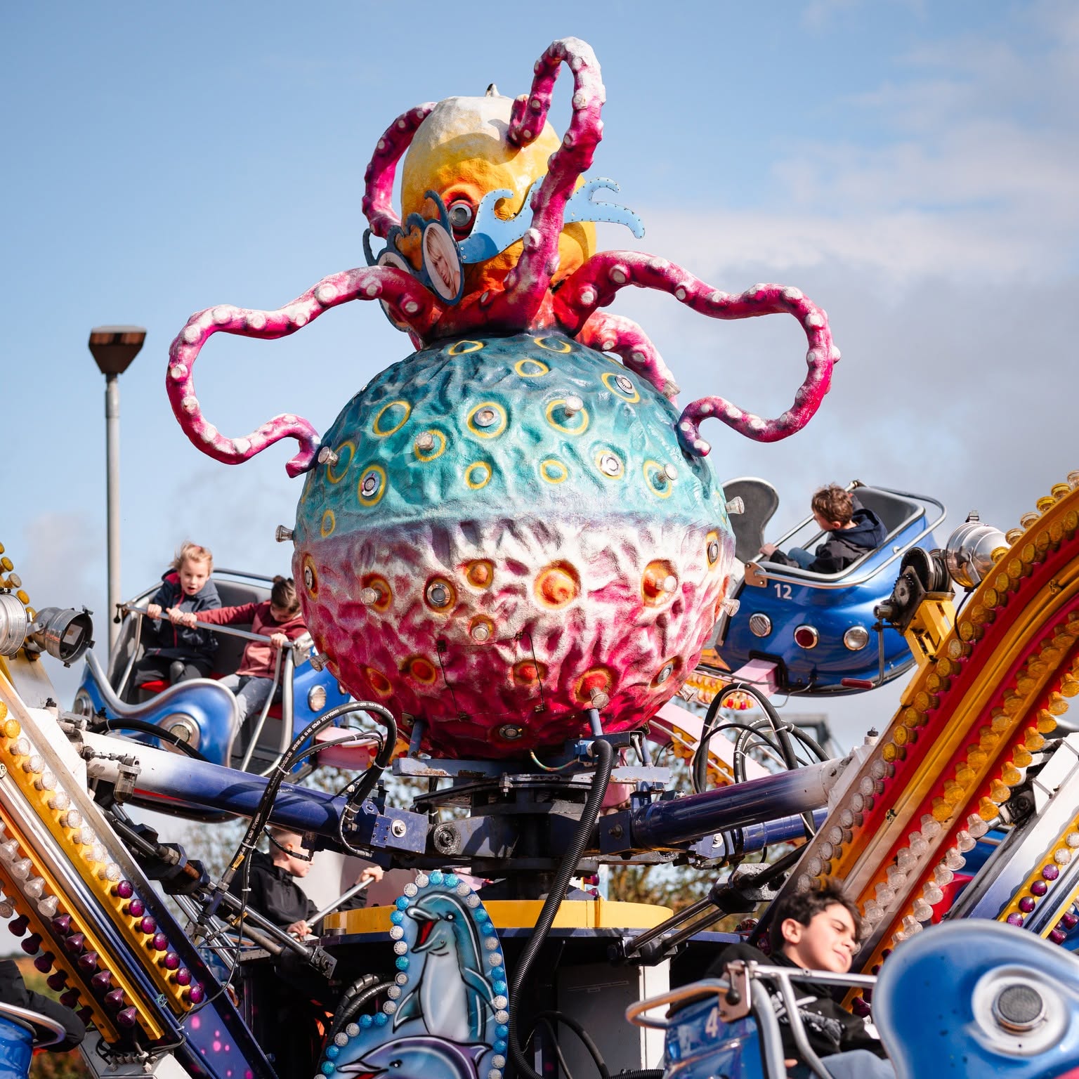 Dieser Alternativtext wurde KI-generiert: Ein farbenfrohes Fahrgeschäft mit einem tentakelartigen, skulpturalen Aufsatz dominiert das Bild. Im Hintergrund sind weitere Fahrgeschäfte und Personen erkennbar.