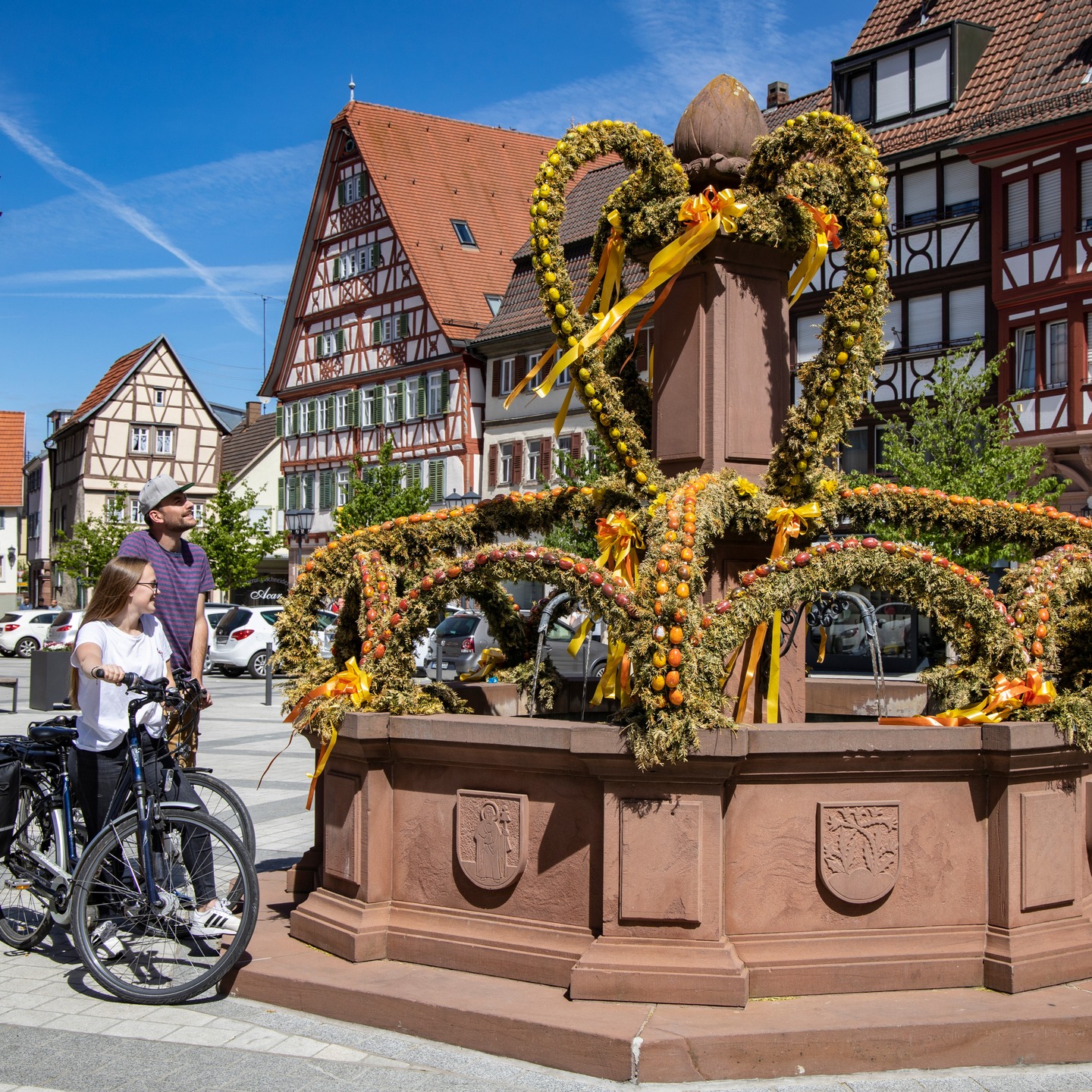 Zwei Radfahrer betrachten einen geschmückten Brunnen auf einem Marktplatz mit Fachwerkhäusern im Hintergrund. Es ist ein sonniger Tag.