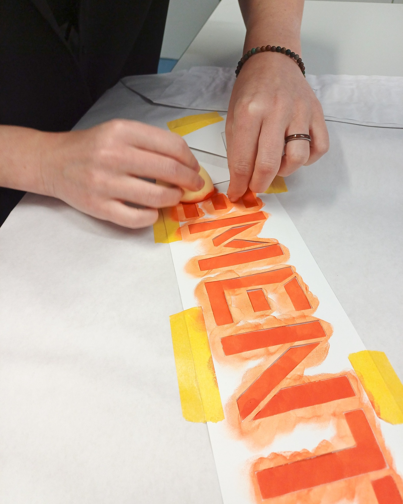 Eine Person entfernt vorsichtig eine Schablone von einem Papier, auf dem mit orangefarbener Farbe ein stilisiertes, geometrisches Muster entstanden ist; gelbe Klebebänder sichern die Schablone am Rand. (KI-Alternativtext)