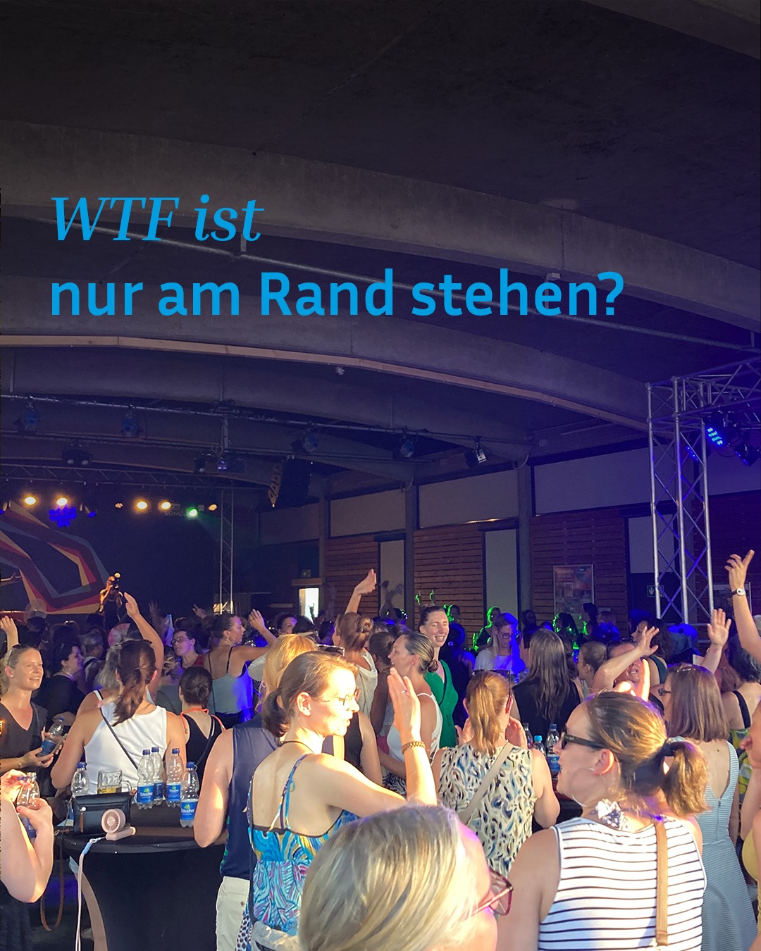 Oben steht in blauer Schrift der Text "WTF ist nur am Rand stehen?". Darunter zeigt sich eine große Menschenmenge bei einer belebten Veranstaltung, bei der viele Personen dicht beieinanderstehen, einige die Hände heben oder Getränke halten. Im Hintergrund ist eine Bühne mit buntem Licht und einer Metallkonstruktion zu erkennen. (KI-Alternativtext)
