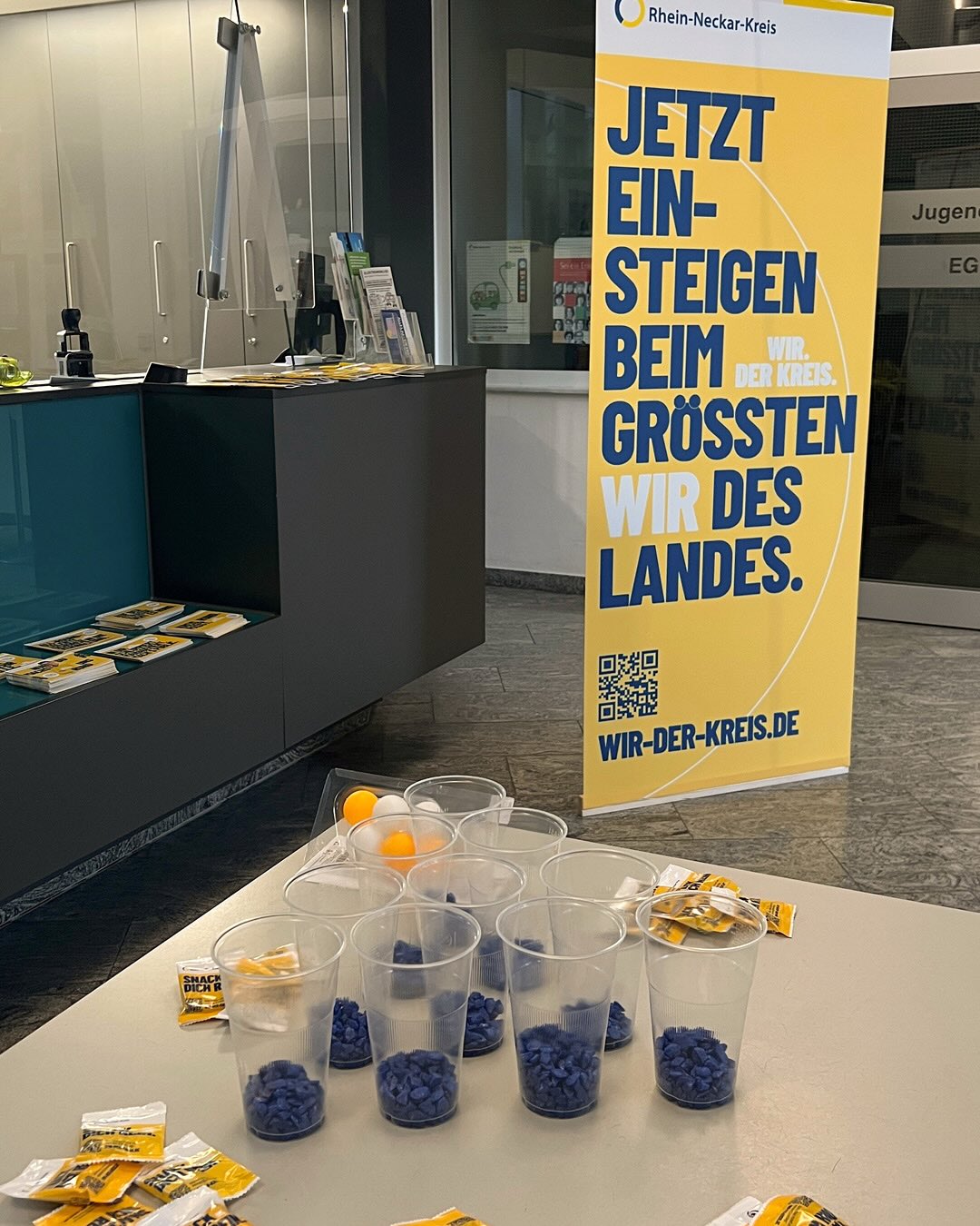Dieser Alternativtext wurde KI-generiert: Ein Tisch mit kleinen, gefüllten Beuteln und einem Ständer mit Informationsmaterialien im Hintergrund. Ein Banner mit Text und einem QR-Code ist ebenfalls zu sehen.