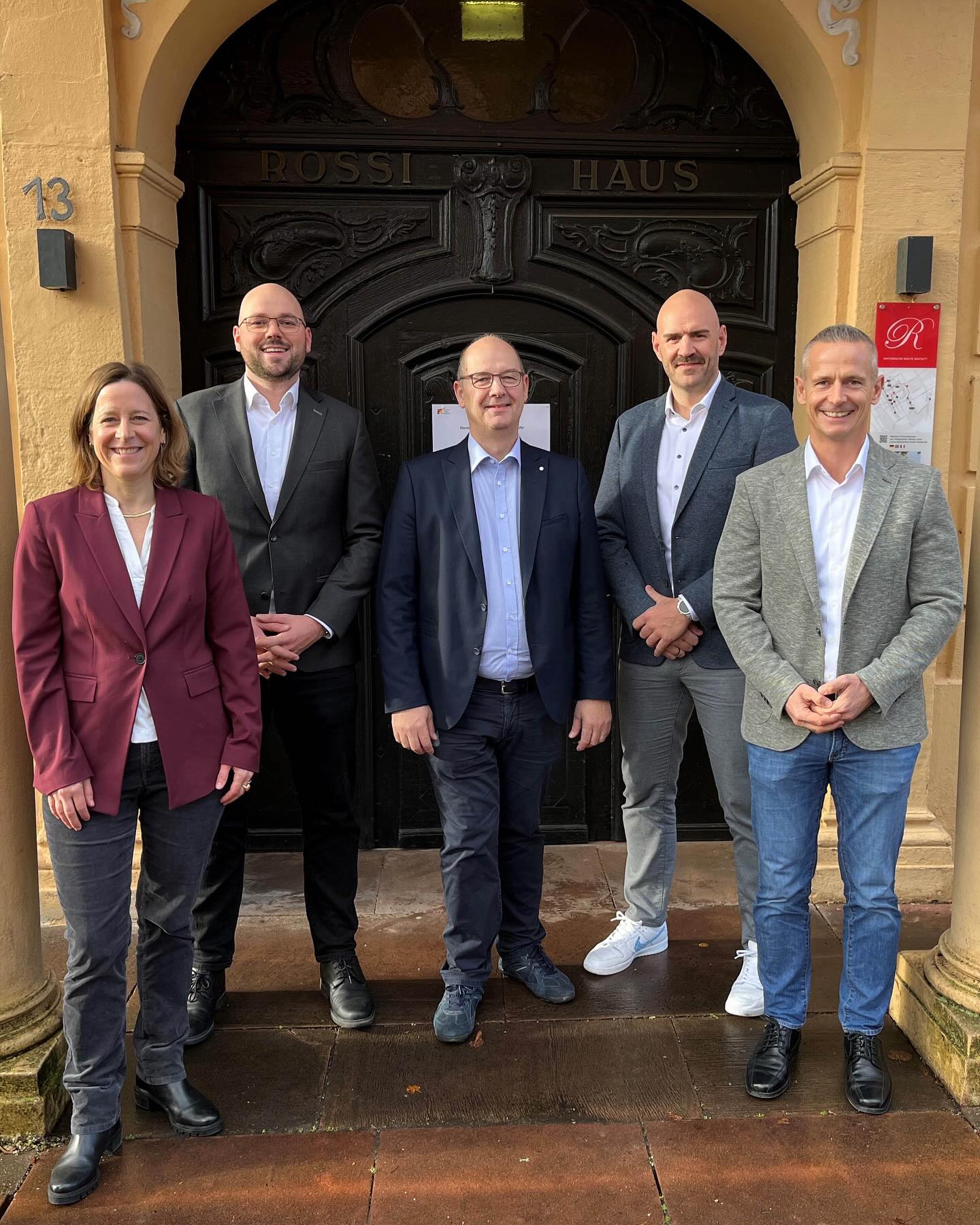 Dieser Alternativtext wurde KI-generiert: Eine Gruppe von vier Personen steht vor einer verzierten Tür. Über der Tür ist ein Schild mit der Aufschrift "ROSSI HAUS" zu sehen.