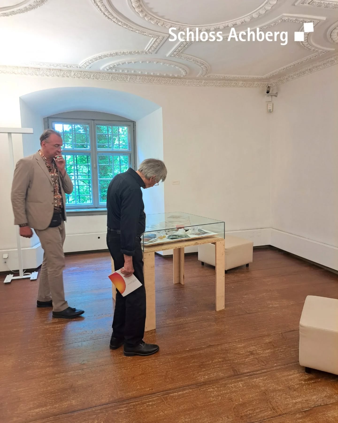 Zwei Personen betrachten eine Vitrine in einem Raum mit Holzfußboden und Stuckdecke. Eine Person hält ein Dokument in der Hand.