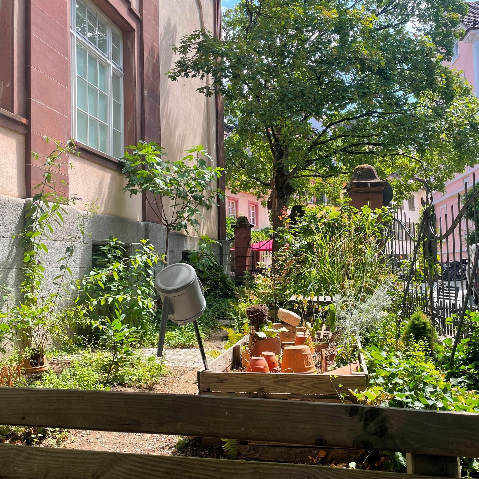 Ein kleiner Gartenbereich mit einem Holzbeet voller umgedrehter Tontöpfe und Pflanzen. Daneben steht eine graue Gießkanne auf einem Ständer vor einer Wand mit roter Fassade und einem Fenster. Im Hintergrund sind ein großer Baum und ein schwarzes Eisengitter zu sehen. (KI-Alternativtext)