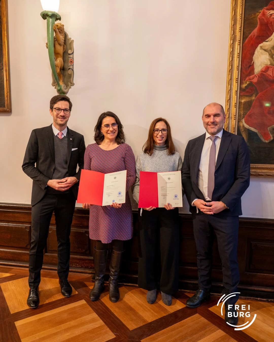 Dieser Alternativtext wurde KI-generiert: Drei Personen stehen nebeneinander und halten jeweils ein rotes Dokument. Die Aufnahme ist in einem Innenraum mit dunkler Holzvertäfelung und einem Gemälde an der Wand.