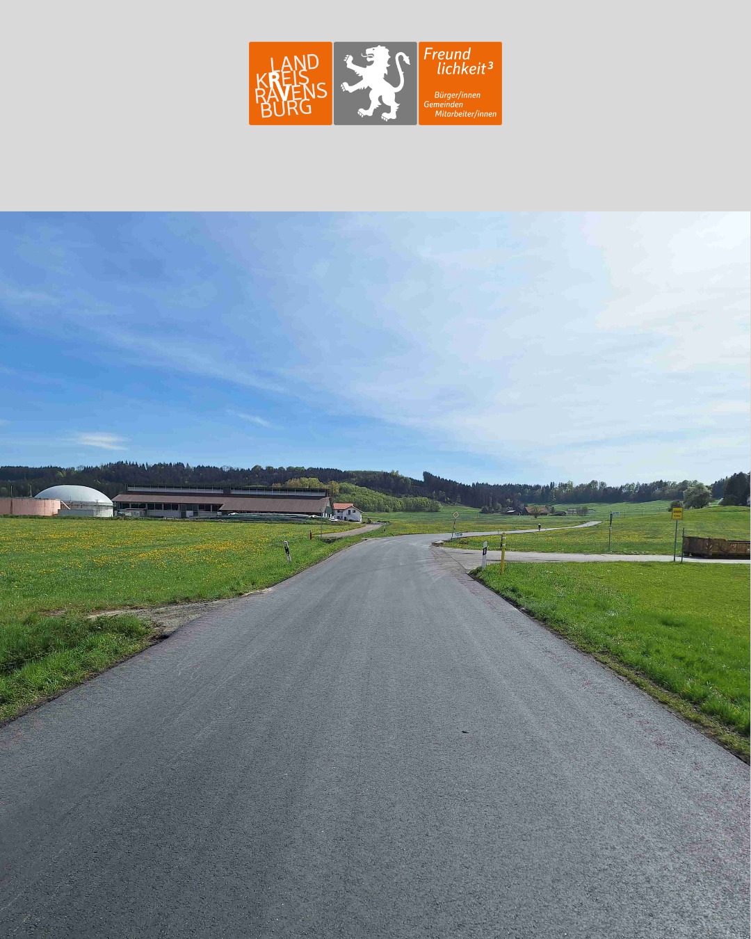 Dieser Alternativtext wurde KI-generiert: Ein farbiges Bild zeigt eine Landstraße, die durch eine grüne Landschaft führt. Im Hintergrund sind Gebäude, Bäume und Hügel zu sehen.