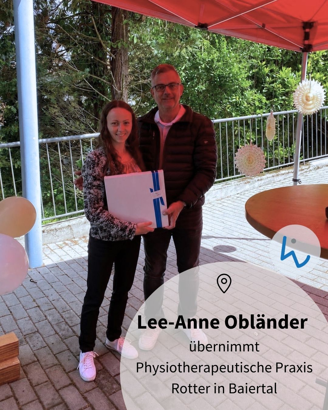 Zwei Personen stehen unter einem roten Zelt und halten gemeinsam ein weißes Dokument mit blauer Binde. Im Vordergrund steht der Text „Lee-Anne Obländer übernimmt Physiotherapeutische Praxis Rotter in Baiertal“. Im Hintergrund sind Bäume, ein Geländer und Dekorationen zu sehen. (KI-Alternativtext)