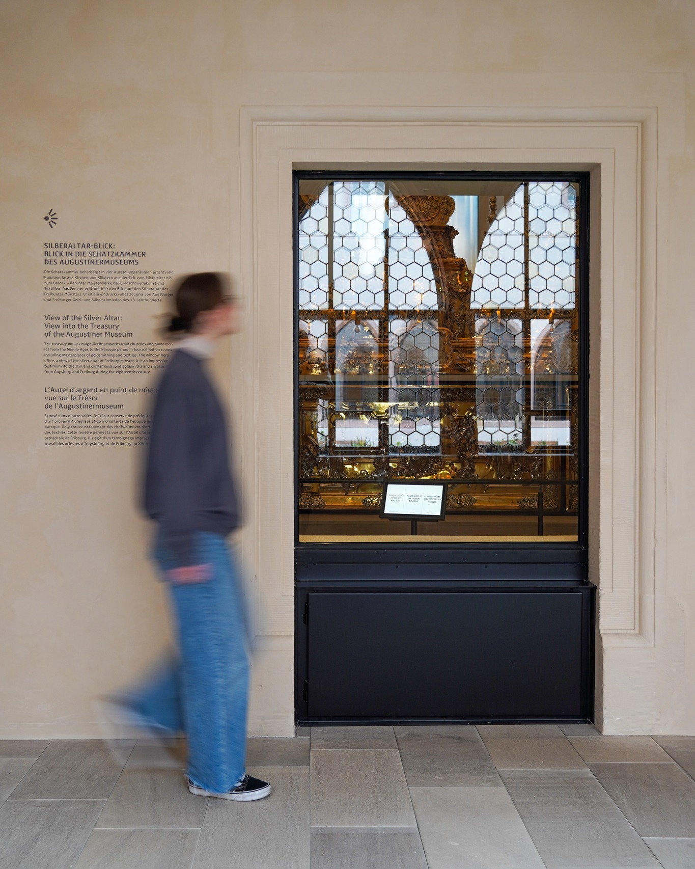Dieser Alternativtext wurde KI-generiert: Ein Innenraum mit einer goldenen Ausstellungsstücke hinter Glas und einem unscharfen Besucher im Vordergrund. Das Bild ist farbig und zeigt Texttafeln in mehreren Sprachen.