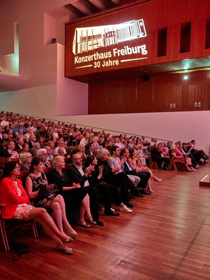 Ein großes Publikum sitzt in einem Saal und applaudiert. An der Wand ist ein Projektionsbild mit der Aufschrift "Konzerthaus Freiburg 30 Jahre" zu sehen. (KI-Alternativtext)