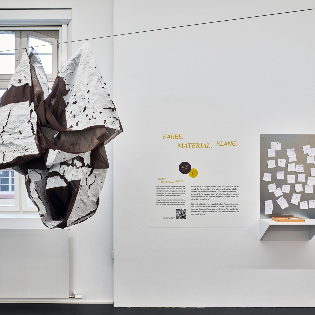 Dieser Alternativtext wurde KI-generiert: Ein Innenraum mit einer farbigen abstrakten Skulptur im Vordergrund. Im Hintergrund befindet sich eine Pinnwand mit Texten und Farbmustern.