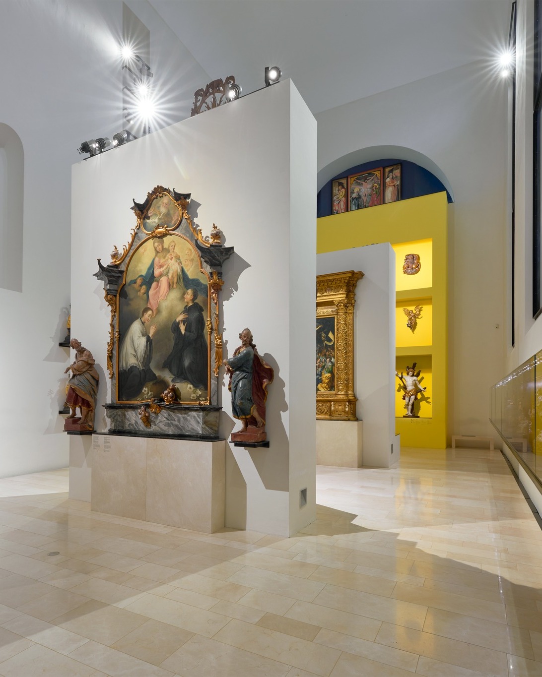 Dieser Alternativtext wurde KI-generiert: Innenaufnahme eines hellen Raumes, wahrscheinlich in einem Museum, mit religiösen Kunstwerken und Skulpturen. Verschiedene Gemälde und Figuren stehen an Wänden und auf Podesten.