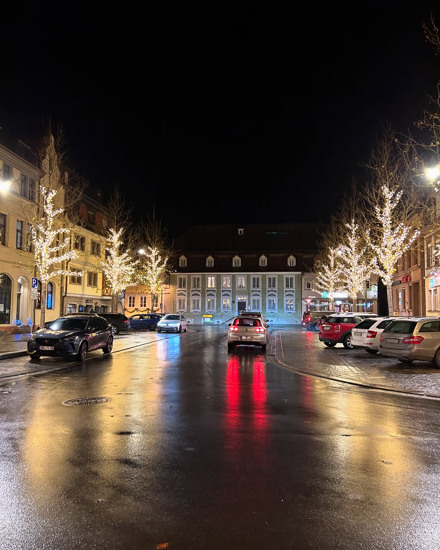 Ein nächtlicher Marktplatz mit festlich geschmückten Bäumen und parkenden Autos. Die nassen Straßen spiegeln die Lichter der Gebäude und Fahrzeuge.