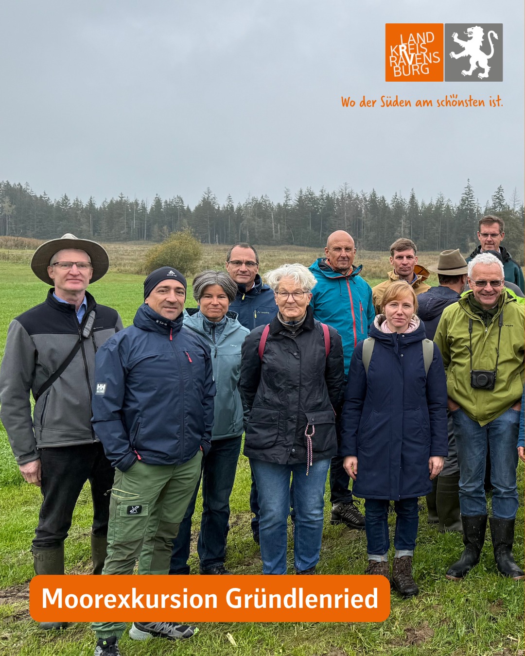Dieser Alternativtext wurde KI-generiert: Eine Gruppe von Personen steht auf einer Wiese vor einer Baumreihe. Im unteren Bildbereich befindet sich ein orangefarbener Balken mit einem Text.