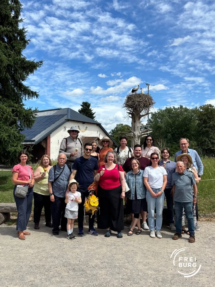 Gruppe von Erwachsenen und einem Kind vor einem Gebäude mit Solarpanelen und einem Storchennest auf einem Baumstumpf im Hintergrund, unter blauem Himmel mit Wolken. Einige halten Eis in der Hand, im Vordergrund ein gelber Rucksack. Rechts unten das Logo „FREIBURG“. (KI-Alternativtext)