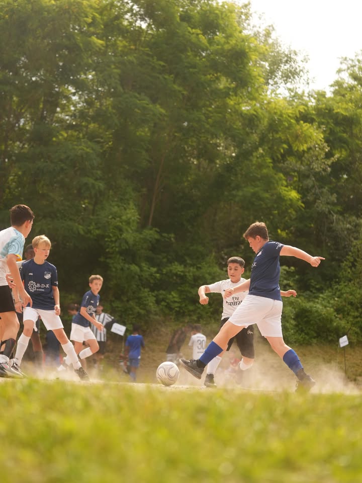 Mehrere Kinder spielen Fußball auf einem Grasfeld vor einem bewaldeten Hintergrund. Ein Kind im blauen Trikot tritt gerade nach dem Ball, während andere Spieler in blauen und weißen Trikots im Spiel sind. Staub wirbelt auf, wo die Füße den Boden berühren. (KI-Alternativtext)