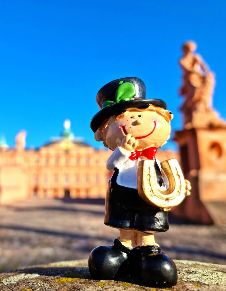 Dieser Alternativtext wurde KI-generiert: Eine kleine Figur mit Hut und Glücksbringer steht im Vordergrund vor einer historischen Stadtlandschaft. Das farbenfrohe Bild fängt einen sonnigen Tag ein.