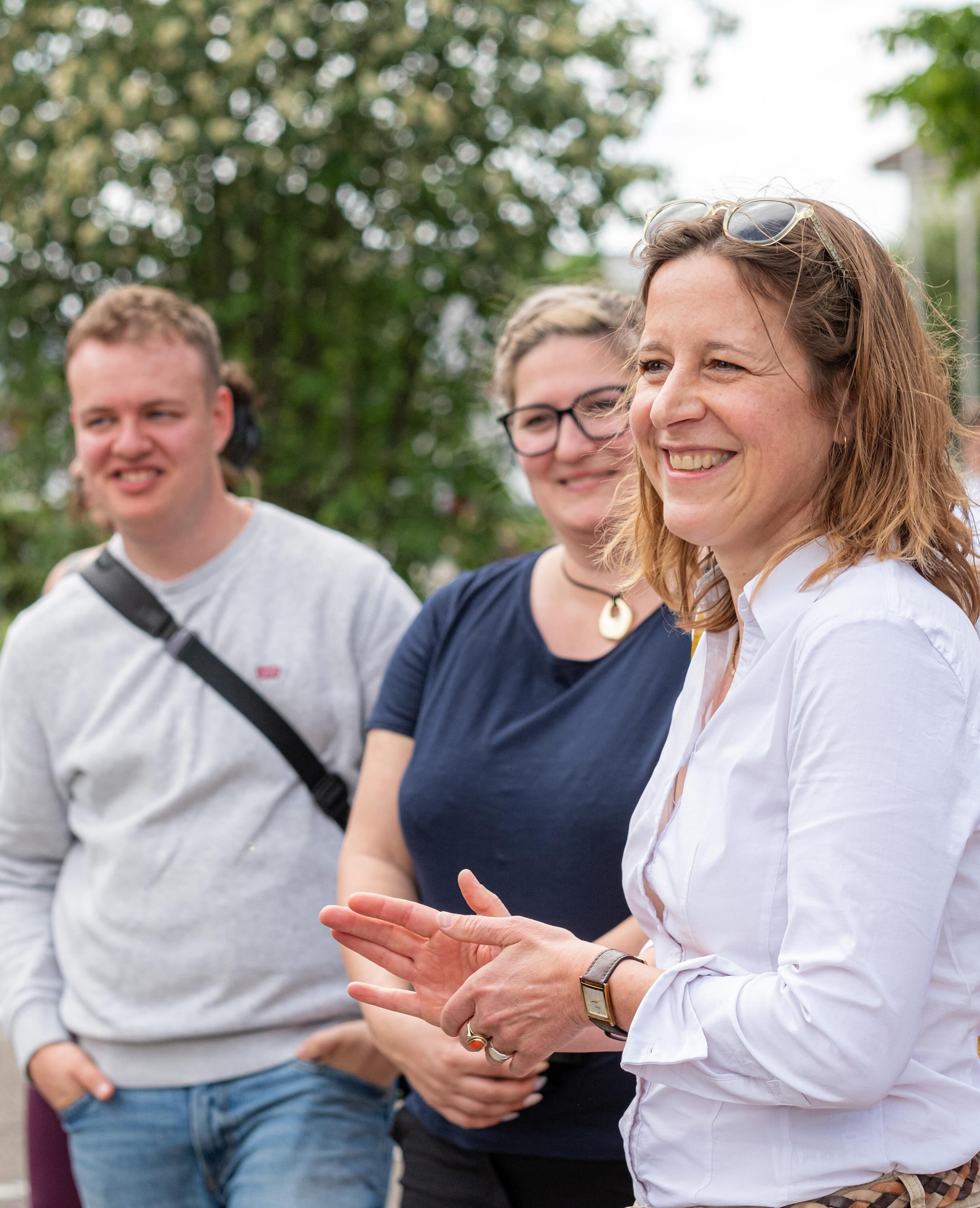 Dieser Alternativtext wurde KI-generiert: Mehrere Personen im Freien, von denen eine lächelt und die Hände hält. Im Hintergrund sind weitere Personen und Gebäude erkennbar.