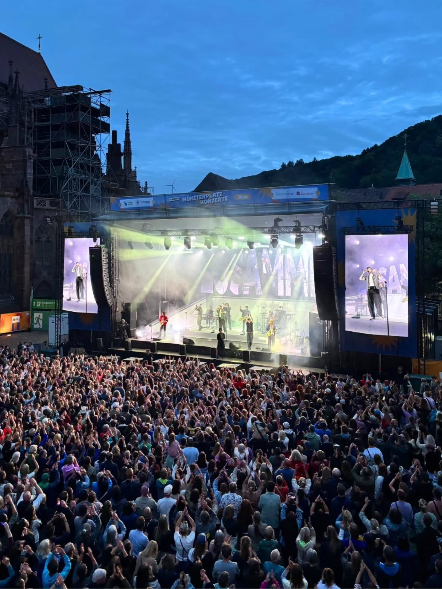 Große Menschenmenge applaudiert auf einem Konzert im Freien vor einer Bühne mit Band und großen Bildschirmen; im Hintergrund gotische Kirche und bewaldeter Hügel bei Dämmerung. Auf der Bühne steht „MÜNSTERPLATZ KONZERTE 2024“. (KI-Alternativtext)