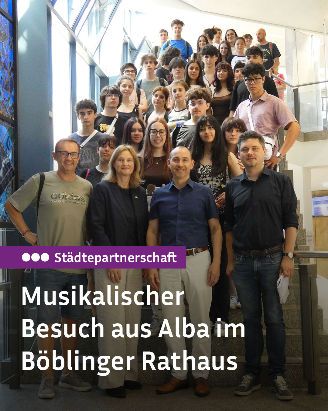 Eine große Gruppe von Personen posiert auf einer Treppe in einem hellen Gebäude. Ein Textfeld im Vordergrund lautet: „Städtepartnerschaft Musikalischer Besuch aus Alba im Böblinger Rathaus“. (KI-Alternativtext)