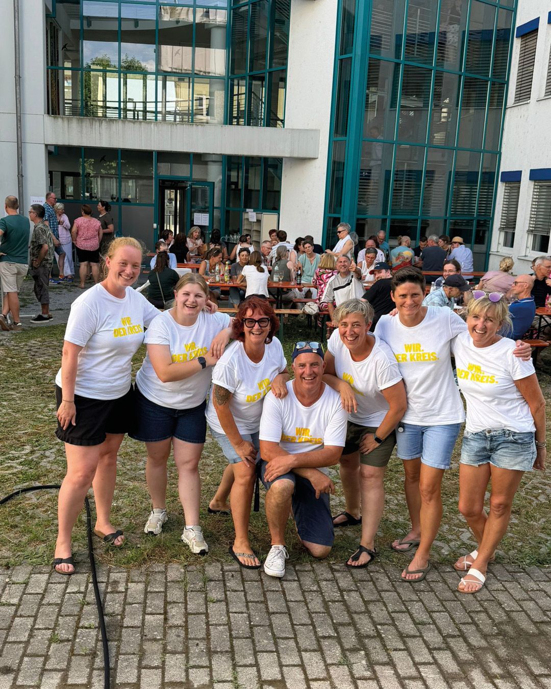 Eine Gruppe von sieben Personen posiert lächelnd im Vordergrund vor einem Gebäude mit Glasfassade. Sie tragen weiße T-Shirts mit der gelben Aufschrift "WIR. DER KREIS." und haben sich gegenseitig umarmt. Im Hintergrund sitzen und stehen viele weitere Personen bei einem geselligen Treffen im Freien. (KI-Alternativtext)