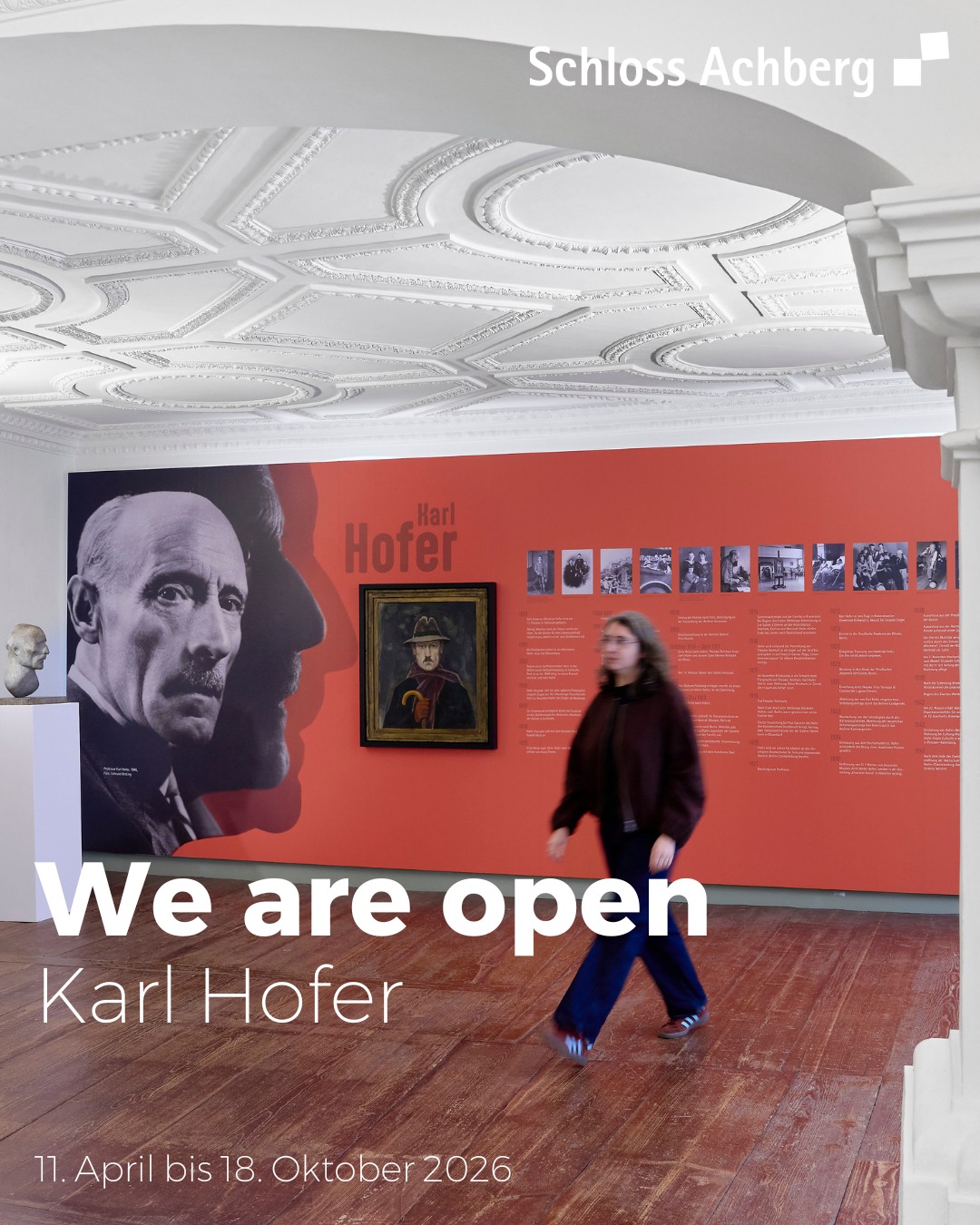 Dieser Alternativtext wurde KI-generiert: Ein Innenraum mit einer verzierten Decke und einer roten Wand mit Ausstellungsstücken und Texten. Eine Person steht vor der Ausstellung und blickt zur Seite.