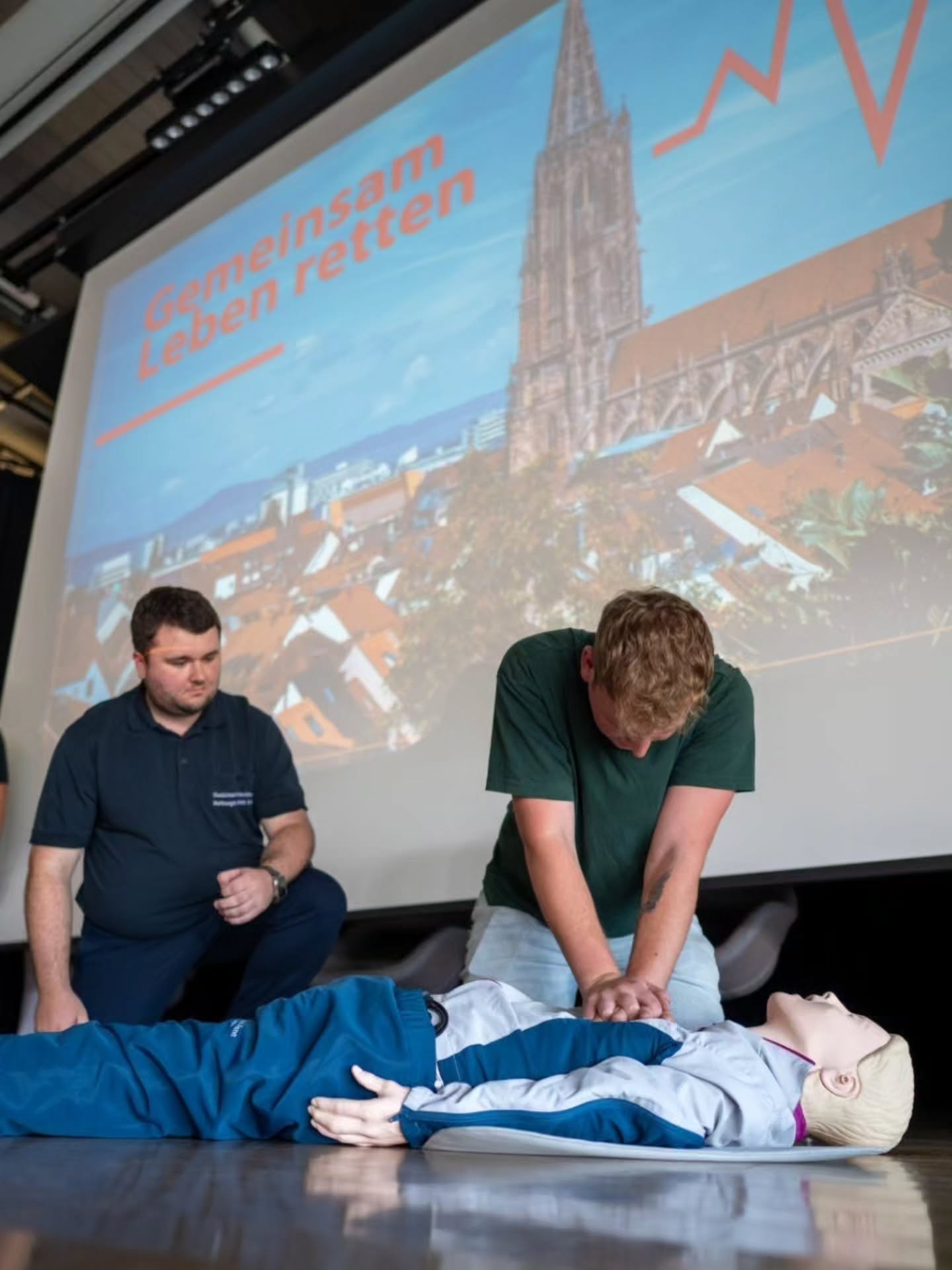Eine Person führt an einer Puppe Reanimation durch, während eine zweite Person daneben kniet und zuschaut. Im Hintergrund ist eine Projektion mit dem Text „Gemeinsam Leben retten“ und einer Kirche zu sehen. (KI-Alternativtext)