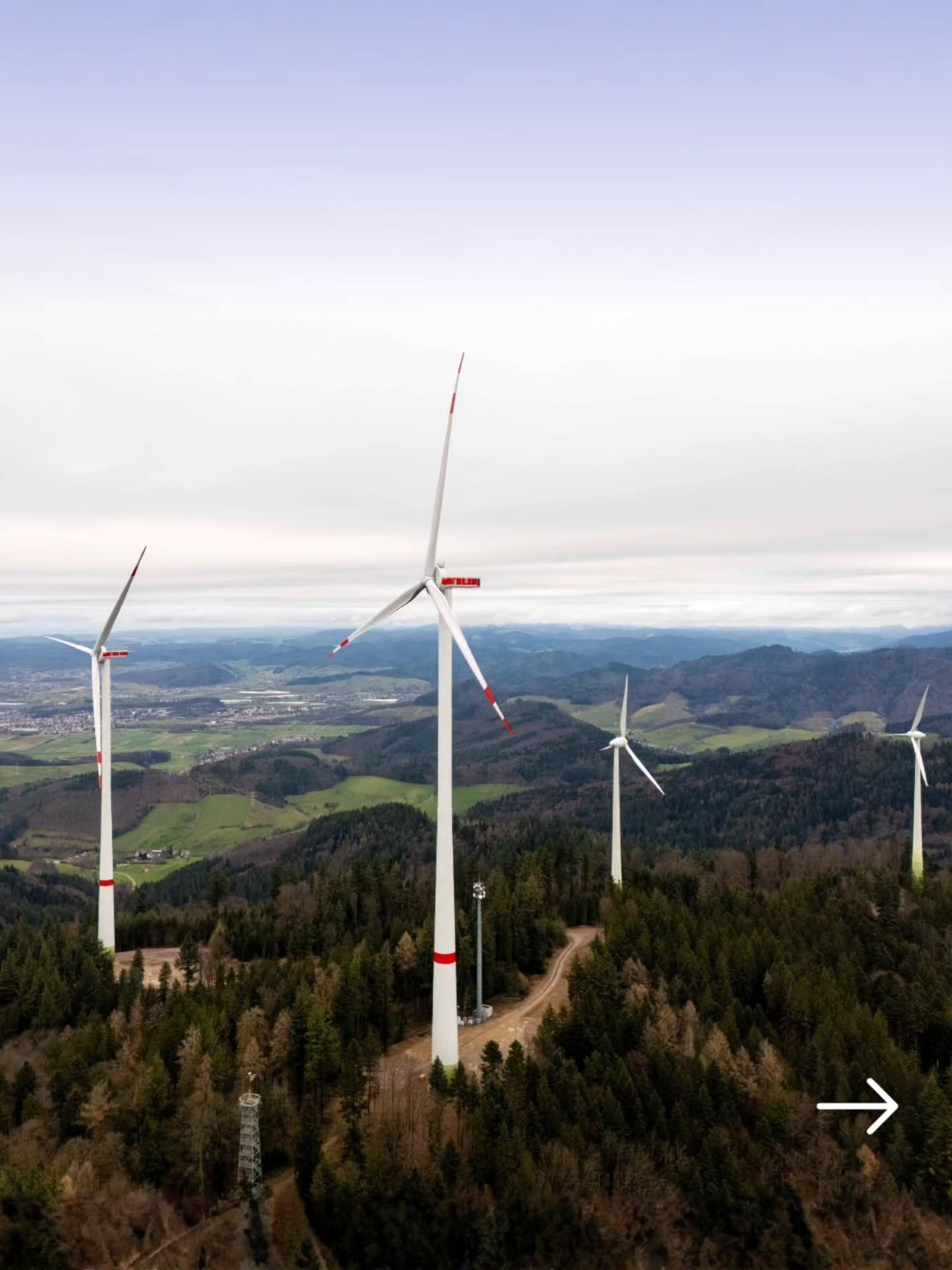 Dieser Alternativtext wurde KI-generiert: Mehrere Windkraftanlagen stehen in einer bewaldeten Hügellandschaft. Im Hintergrund erstreckt sich eine weitläufige Landschaft mit Bergen und Wolken.