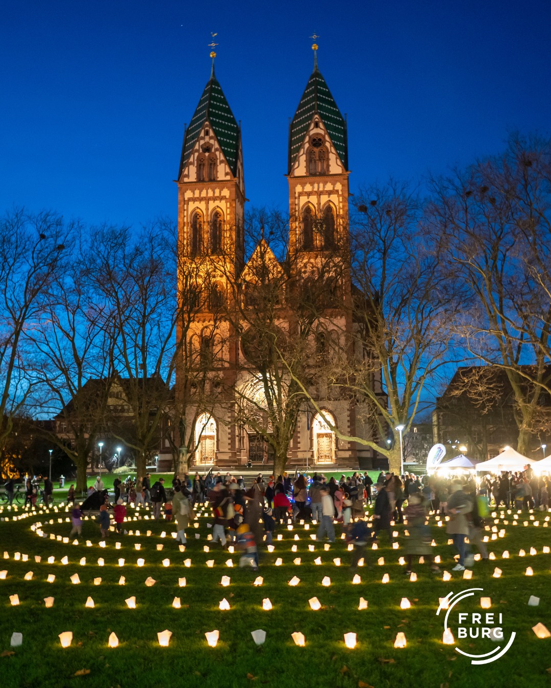 Dieser Alternativtext wurde KI-generiert: Ein hochaufragendes Gebäude mit zwei spitzen Türmen ist im Abendlicht zu sehen. Vor dem Gebäude befindet sich eine große Wiese mit vielen kleinen Lichtern und Menschen.