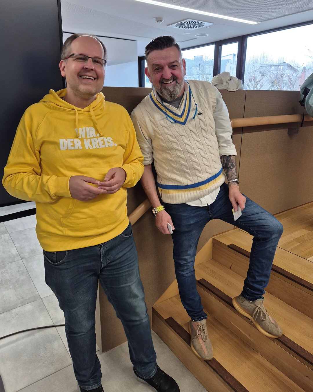 Dieser Alternativtext wurde KI-generiert: Zwei lächelnde Personen stehen nebeneinander auf einer Holztreppe. Beide tragen Jeans und Oberteile, eine Person hat eine gelbe Jacke an.