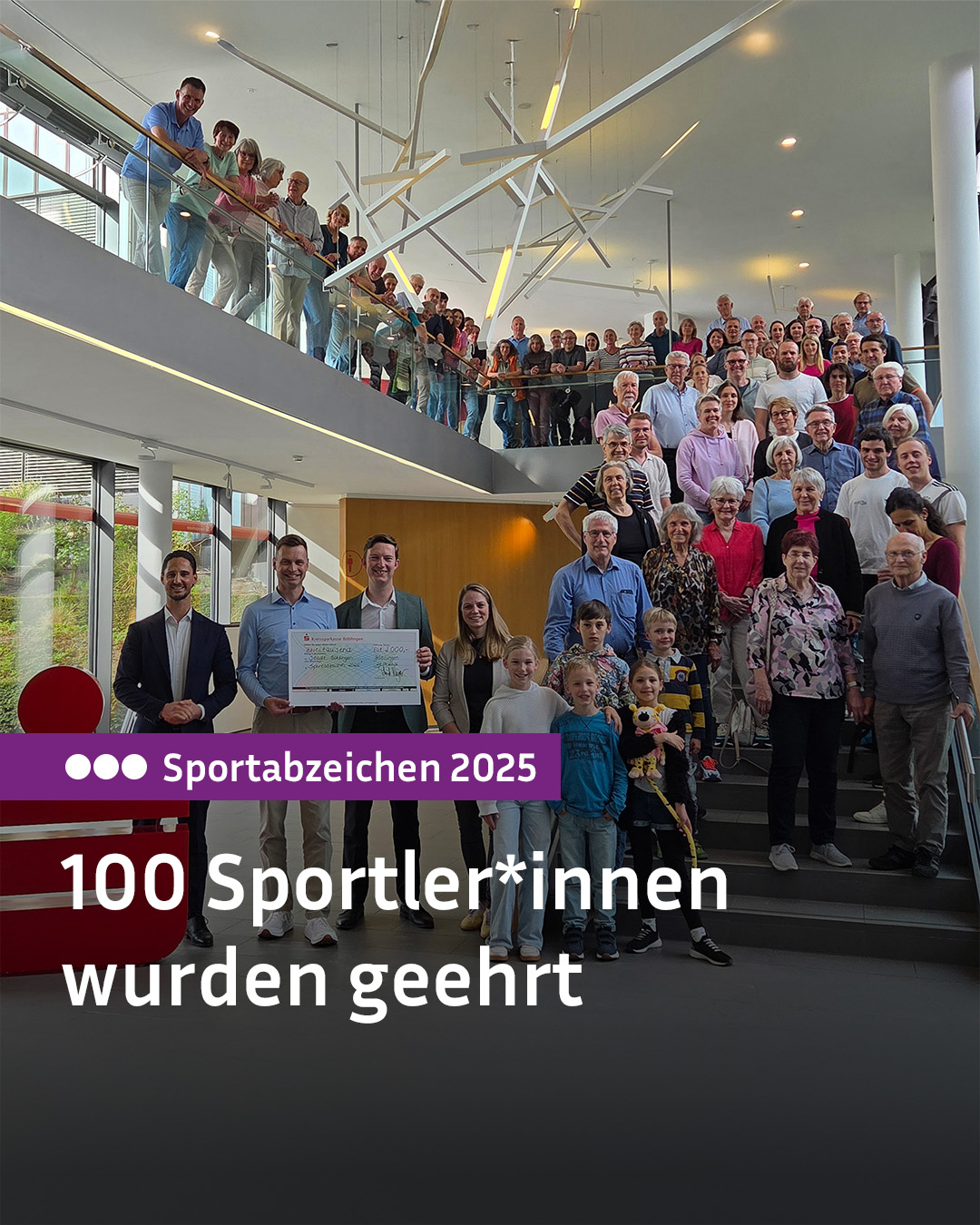 Dieser Alternativtext wurde KI-generiert: Eine große Gruppe von Menschen posiert auf einer Treppe in einem hellen Gebäude. Im Vordergrund hält jemand einen Scheck, und ein Banner mit der Aufschrift „Sportabzeichen 2025“ ist zu sehen.