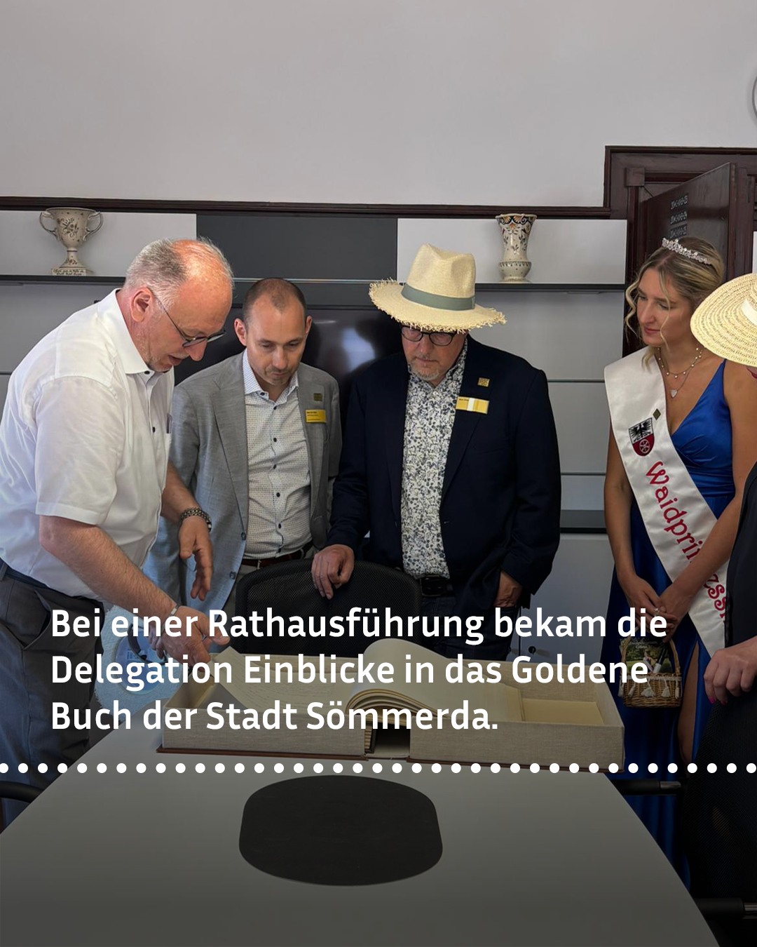 Bei einer Rathausführung betrachtet eine Delegation das Goldene Buch der Stadt Sömmerda. Eine Person in blauem Kleid mit Schärpe und Kopfbedeckung steht daneben. Im Hintergrund sind Regale mit Dekorationsgegenständen zu sehen. (KI-Alternativtext)