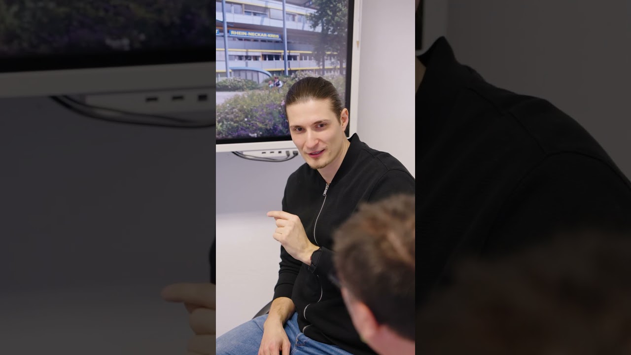 Dieser Alternativtext wurde KI-generiert: Eine Person sitzt und schaut in die Kamera, während im Hintergrund ein Gebäude zu sehen ist. Das Bild ist farbig und zeigt eine Innenansicht.