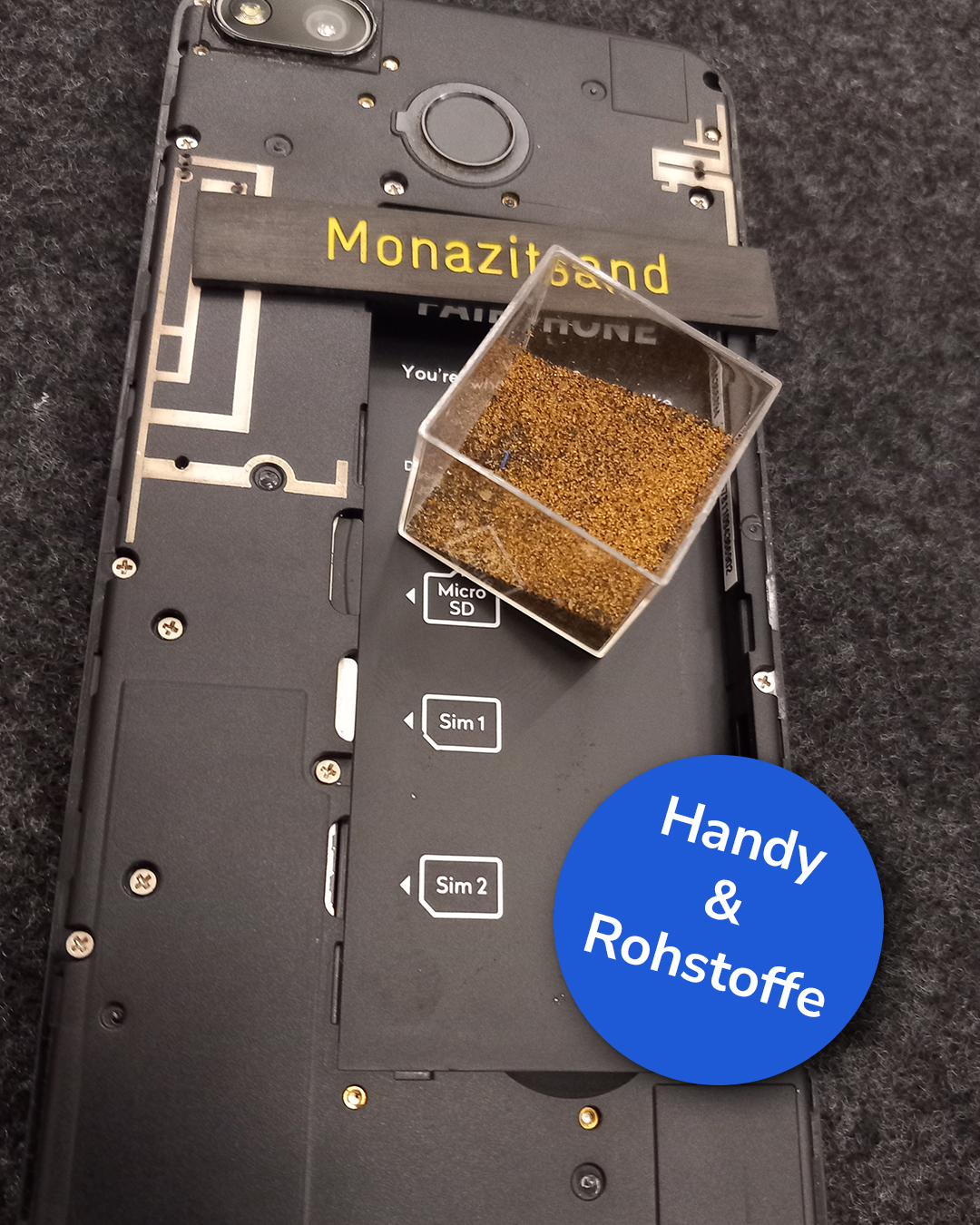 Dieser Alternativtext wurde KI-generiert: Eine Nahaufnahme zeigt die Rückseite eines Mobiltelefons und ein kleines Gefäß mit braunem Sand. Beschriftungen und Symbole sind auf dem Gerät und einem separaten Stück Papier zu sehen.