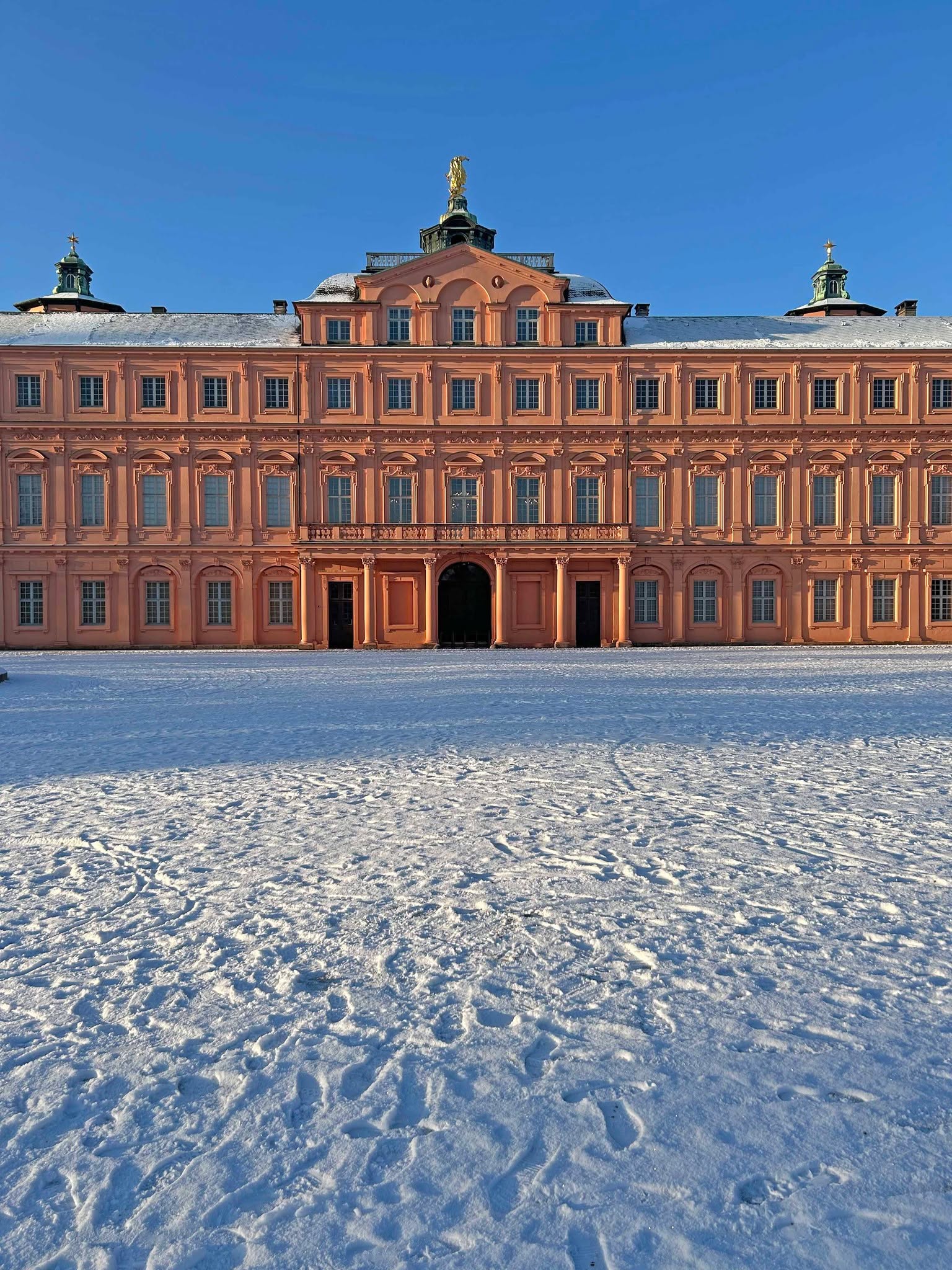 Dieser Alternativtext wurde KI-generiert: Ein rotes Gebäude mit vielen Fenstern steht vor einer schneebedeckten Fläche. Es ist ein klarer, blauer Himmel zu sehen.