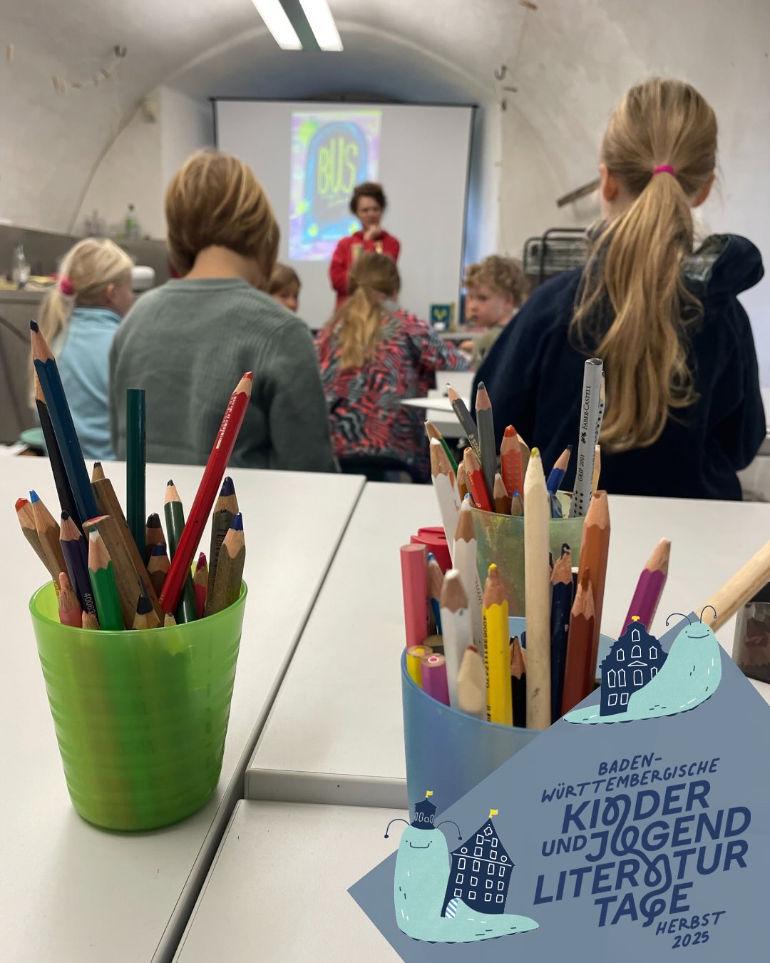 Dieser Alternativtext wurde KI-generiert: Ein Innenraum mit einer Gruppe von Personen, die einer Präsentation zuwenden. Auf einem Tisch befinden sich viele Buntstifte und Werbematerialien.