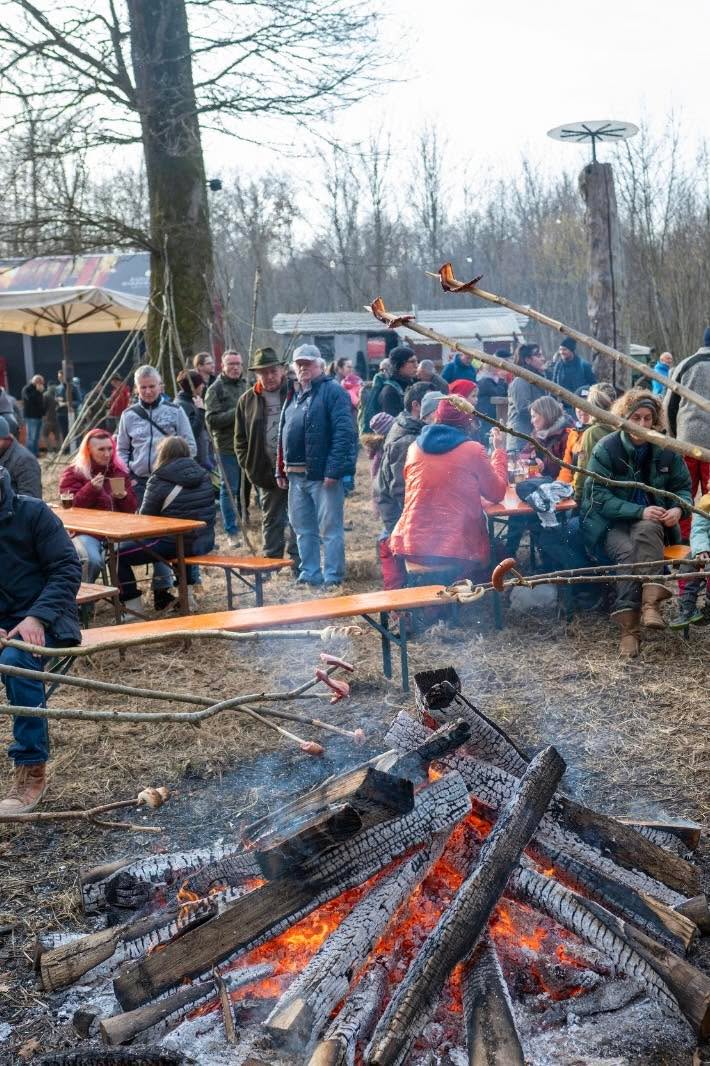 Dieser Alternativtext wurde KI-generiert: Eine Gruppe von Menschen versammelt sich im Freien um ein großes Lagerfeuer. Es werden Speisen über dem Feuer zubereitet und gegessen, während andere an Tischen sitzen.