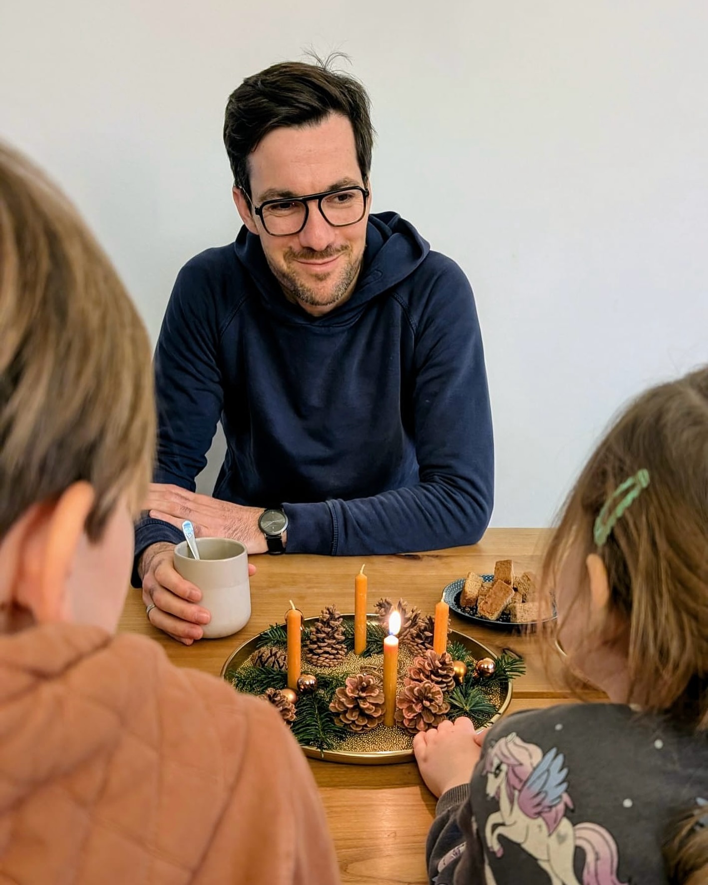 Dieser Alternativtext wurde KI-generiert: Eine Person mit Brille sitzt am Tisch und blickt in die Kamera. Auf dem Tisch befinden sich Kerzen, Dekoration und Essen.