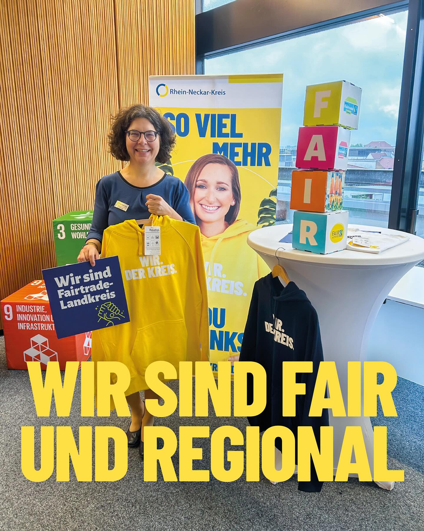 Dieser Alternativtext wurde KI-generiert: Eine Person steht vor einem Werbestand mit Bannern und Plakaten. Im Hintergrund ist ein Fenster mit Blick auf eine Stadt zu sehen.