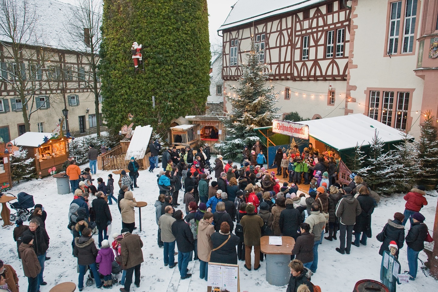 Ein Weihnachtsmarkt mit vielen Menschen, Buden und geschmückten Tannenbäumen. Es liegt Schnee auf dem Boden.