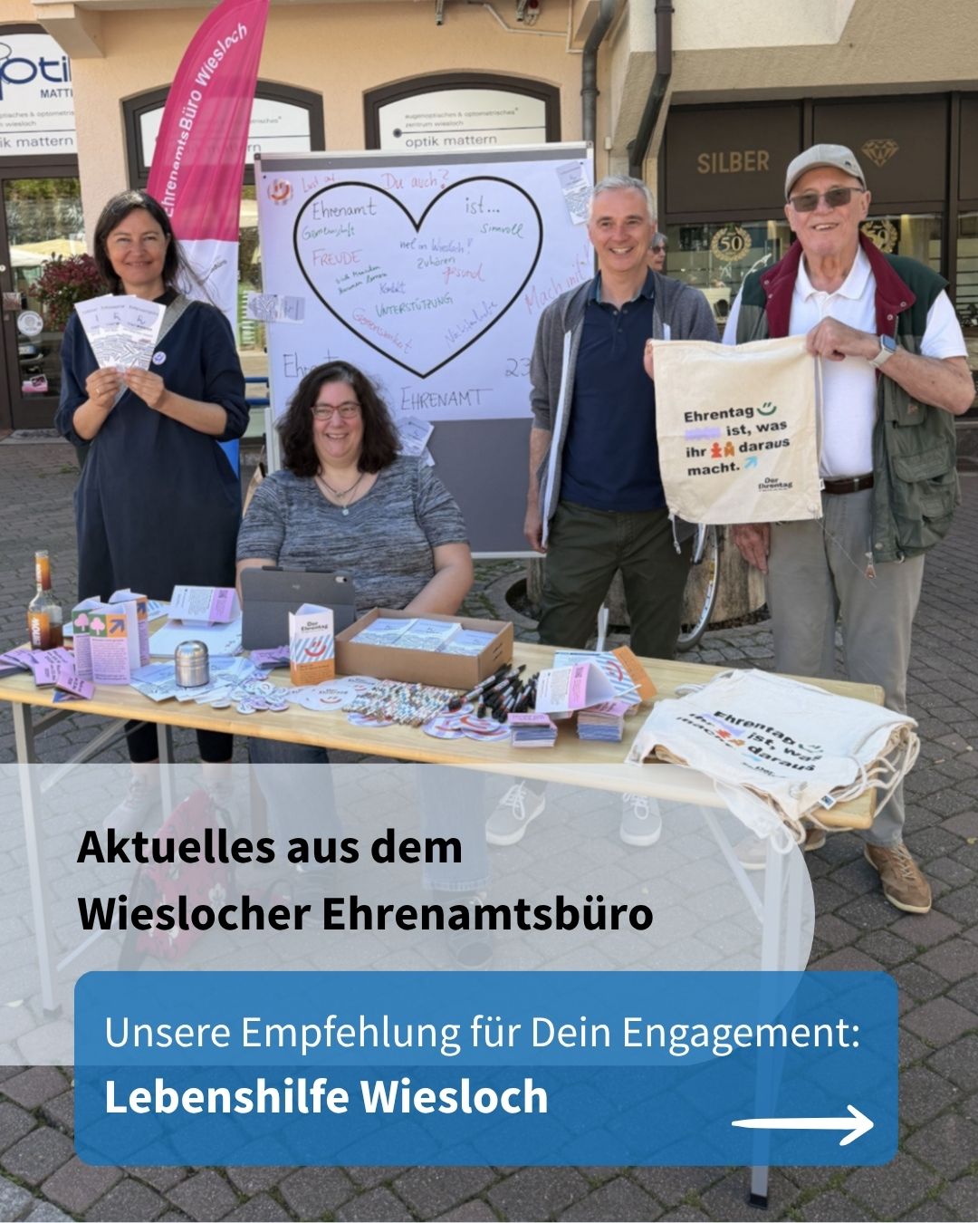 Vier Personen stehen an einem Informationsstand im Freien, einer hält eine Tasche mit der Aufschrift „Ehrentag ist, was ihr daraus macht.“ Im Hintergrund ein Whiteboard mit Herz und Stichworten zum Ehrenamt sowie ein Banner des Ehrenamtsbüros Wiesloch. Unten Text: „Aktuelles aus dem Wieslocher Ehrenamtsbüro – Unsere Empfehlung für Dein Engagement: Lebenshilfe Wiesloch“. (KI-Alternativtext)