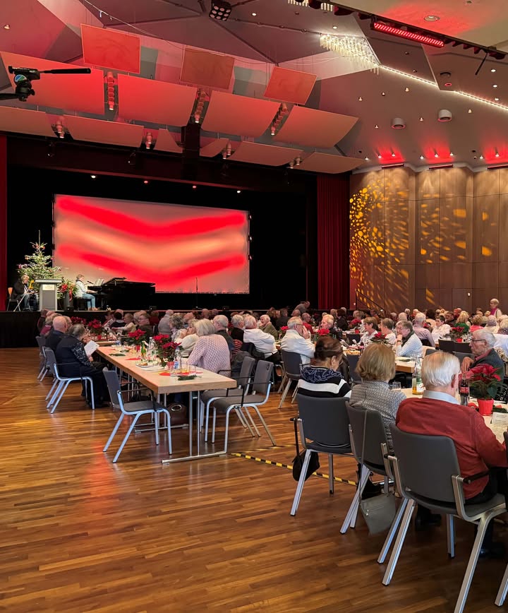 Dieser Alternativtext wurde KI-generiert: Ein großer Raum mit Tischen und Stühlen, an denen Menschen sitzen. Im Hintergrund befindet sich eine Bühne mit einem großen Bildschirm, der rot beleuchtet ist.